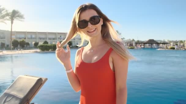 Retrato de menina sorrindo, olhando para a câmera. Mulher bonita em fato de banho vermelho e óculos de sol perto da piscina no dia ensolarado quente. Menina se divertindo, férias de verão na villa de luxo . — Vídeo de Stock