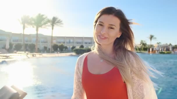 Aantrekkelijk jong meisje met gouden haren waait in de wind, in rode sexy bikini, draait om de camera en glimlacht. Zwembad strand achtergrond. Vrouw zonnen en ontspannen in het Resort. Zomervakantie. — Stockvideo