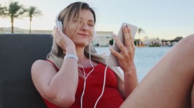 Kırmızı bikini beyaz kulaklık otel havuz yan sandalyede güverte üzerinde yatarken, onun smartphone üzerinden müzik dinleme gülümseyen genç çekici genç kadın portresi. Kız rahatlatıcı Resort