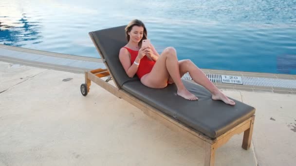 时尚性感的女人穿着红色比基尼躺在甲板上的椅子上, 使用智能手机应用程序, 浏览互联网, 在酒店泳池边区域发短信的最高视野。女孩博客在线和日光浴, 在度假村放松. — 图库视频影像
