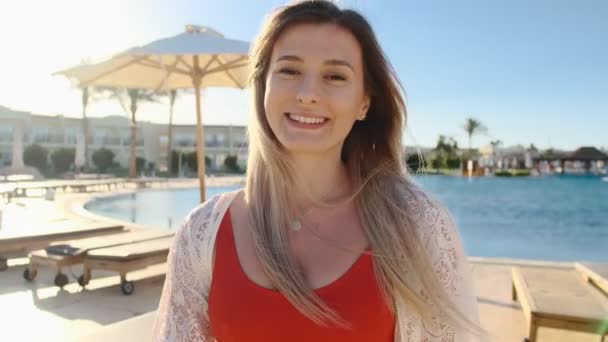 Smiling young girl, looking at camera. Beautiful woman in red swimsuit near the blue swimming pool on hot sunny day. Girl having good time, summer holiday at luxury villa. — Stock Video