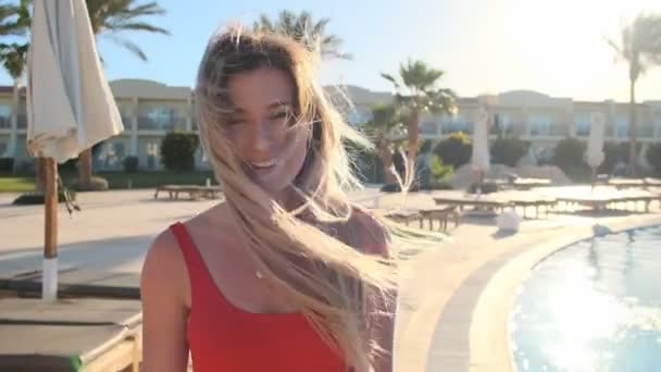 Portrait of charming young girl with golden hair blowing in wind, in red sexy bikini, turns to camera and smiles. Pool beach background. Woman sunbathing and relaxing at resort. Summer holidays — Stock Video