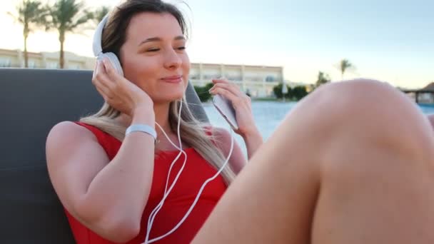 Ritratto di giovane donna attraente sorridente in bikini rosso e cuffie bianche che ascolta musica dal suo smartphone, canta e balla sdraiata sulla sedia a sdraio nell'area laterale della piscina dell'hotel . — Video Stock