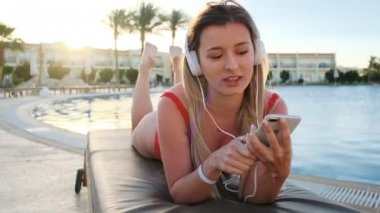 Otel havuzu yan alanında güverte sandalyede yatarken kırmızı bikini ve beyaz kulaklıklar şarkı ve dans gülümseyen genç çekici genç kadın portre. Kız güneşlenme ve tesiste rahatlatıcı.