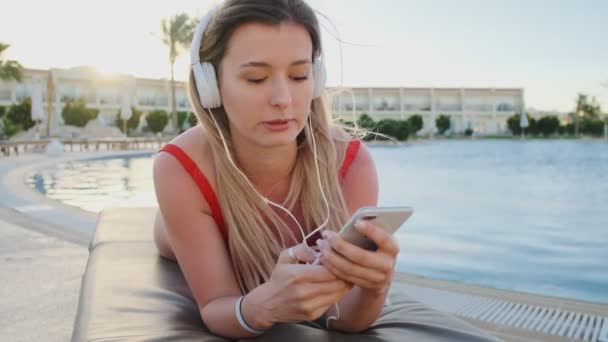 Joven mujer atractiva en bikini rojo y auriculares blancos escuchando música desde su teléfono inteligente, cantando canciones mientras está tumbada en la tumbona en la zona de la piscina del hotel. Chica tomando el sol y relajarse en el complejo . — Vídeo de stock