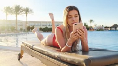 Kadın browsing belgili tanımlık enterne, smartphone, kullanarak bir şezlong üzerinde dinlenme portre sosyal medya aracılığıyla kayar. Havuz kenarında güneşlenirken iken cep telefonunuzu kullanarak kırmızı bikini giyen genç mutlu kız.
