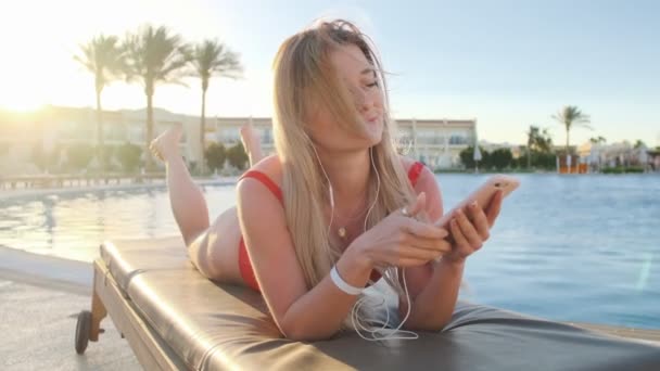 Mulher loira atraente em roupa de banho vermelha dançando, ouvindo música de seu smartphone, enquanto deitada na cadeira de praia na área lateral da piscina do hotel. Menina banhos de sol e relaxante no resort . — Vídeo de Stock