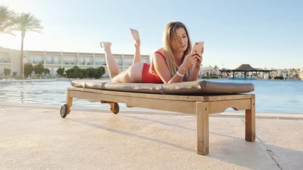 Симпатична блондинка в червоному купальнику переглядає Інтернет на смартфоні, лежачи на стільці біля бокової зони басейну. Розслаблена дівчина смс-повідомлення та блог з мобільним телефоном у відпустці . — стокове відео