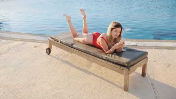 Top view of stylish sexy woman in red bikini lying on deck chair, using smartphone app, browsing the internet, texting in hotel pool side area. Girl blogging online and sunbathing, relaxing at resort. — Stock Video