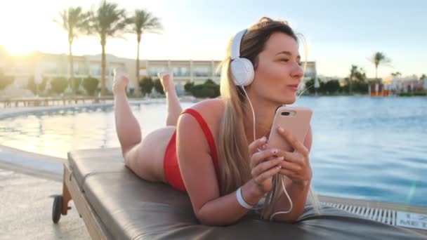 Menina bonito em biquíni vermelho e fones de ouvido brancos ouvir música de seu smartphone, cantando música enquanto deitado na cadeira de praia na área lateral da piscina do hotel. Mulher tomando banho de sol e relaxando no resort . — Vídeo de Stock