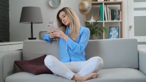 Gyönyörű lány segítségével a digitális mobil készülék böngészés az interneten és a szociális média, tartózkodó kapcsolódik otthon élvezi a modern életmód. Nő segítségével okostelefon a kanapén, textil, míg pihentető. — Stock videók