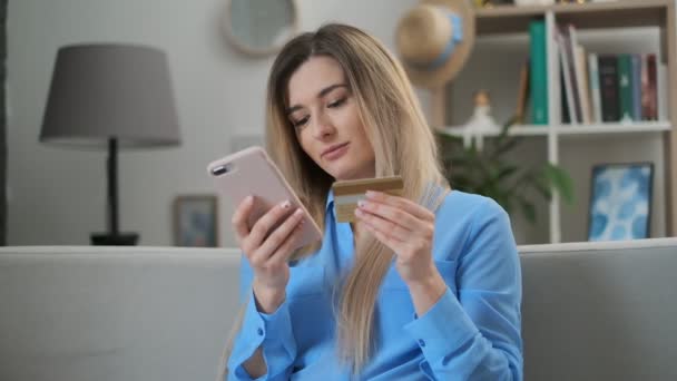 Ragazza compra regali online a casa con carta e telefono app. La donna si siede sul divano, tenendo il numero di carta dello smartphone. Facile transazione di e-commerce, prenotazione, sparatutto online . — Video Stock