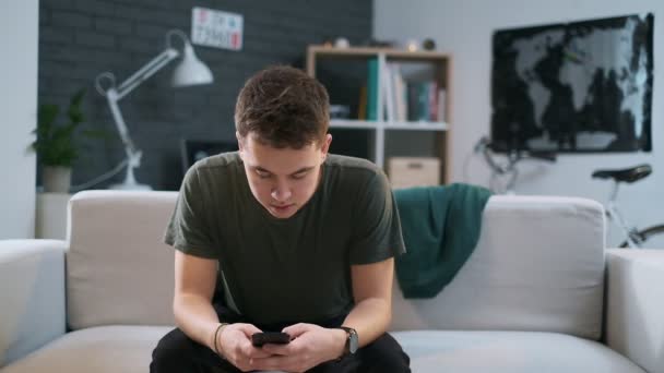 Handsome teenage boy is browsing the web on his phone and then rests back on the couch. — Stock Video