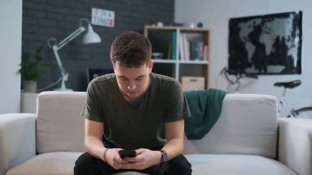 Bonito adolescente está navegando na web em seu telefone e depois descansa de volta no sofá — Vídeo de Stock