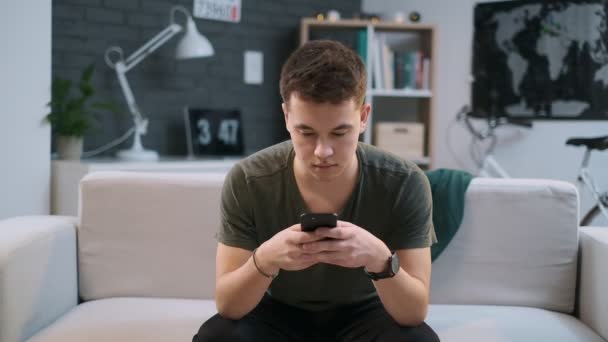 Boy leest een blog op een bank en toont vervolgens het scherm van zijn telefoon naar de camera. — Stockvideo