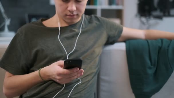 Nahaufnahme, langgezogene Ansicht eines netten Kerls, der auf einer Couch im Raum sitzt, Musik hört und auf seinem Handy blättert — Stockvideo