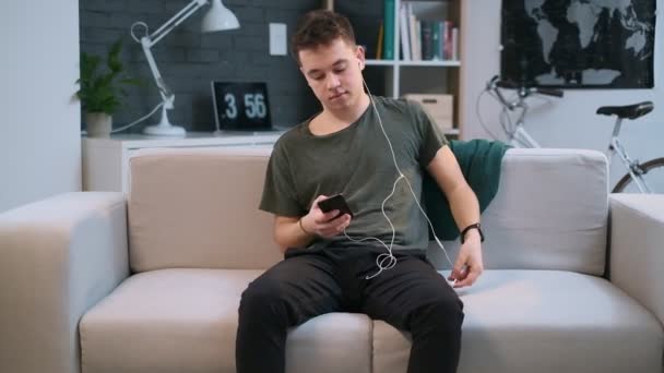 Un adolescente sentado en un sofá está escribiendo en su teléfono celular y usando auriculares al mismo tiempo — Vídeos de Stock