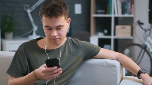 Roztomilá chlapeček poslouchá hudbu ve sluchátkách a používá aplikaci na telefonu, zatímco odpočívá na gauči — Stock video