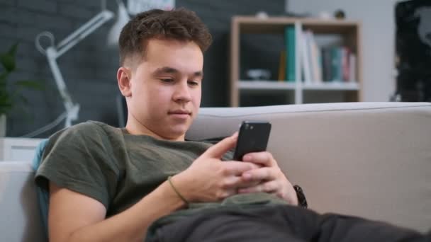Close-up προβολή ενός νεαρού αγοριού κύλιση της τροφοδοσίας σε μια εφαρμογή στο κινητό του τηλέφωνο, ενώ βρίσκεται σε έναν καναπέ στο δωμάτιό του — Αρχείο Βίντεο