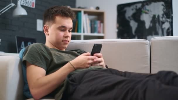 Vista lateral de un niño acostado en un sofá y desplazando la alimentación en su teléfono celular, primer plano — Vídeo de stock