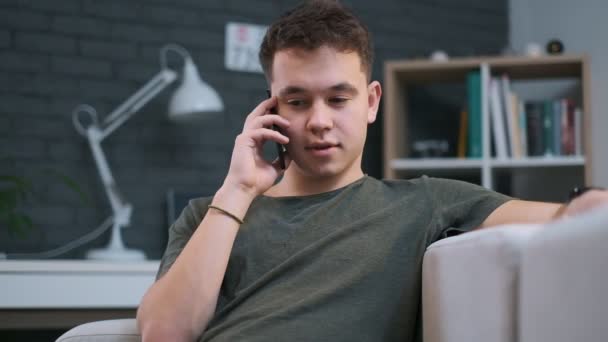 Retrato de cerca de un joven guapo hablando por teléfono y riendo en el sofá de su habitación — Vídeos de Stock