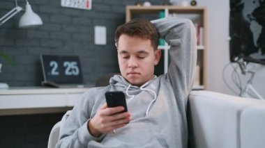 Kapüşonlu bir çocuk kanepede rahatça yatıyor ve evde akıllı telefonuna mesaj atıyor.