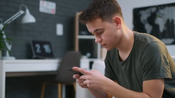 Kant close-up van een tiener geconcentreerd op zijn mobiele telefoon terwijl zittend op de Bank in een stijlvolle kamer — Stockvideo