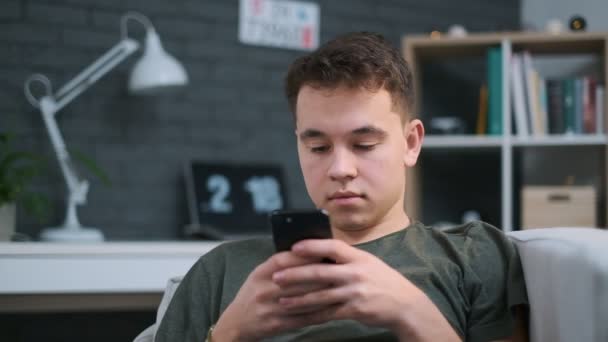 Közeli, kerek portré kilátás egy tinédzser ül a kanapén, és egy app-ra-a telefon — Stock videók