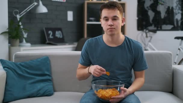 Vista frontal de un adolescente comiendo papas fritas y viendo algo con atención en un sofá — Vídeo de stock