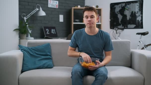 Frontansicht eines hübschen Jungen, der in seiner Freizeit Chips auf einer Couch zu Hause kaut — Stockvideo