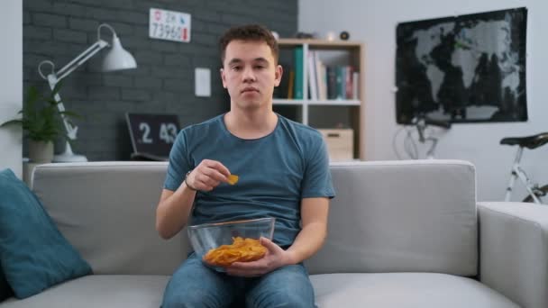 Frontansicht eines gelangweilten Teenagers, der auf einem Sofa sitzt und zu Hause Chips isst — Stockvideo