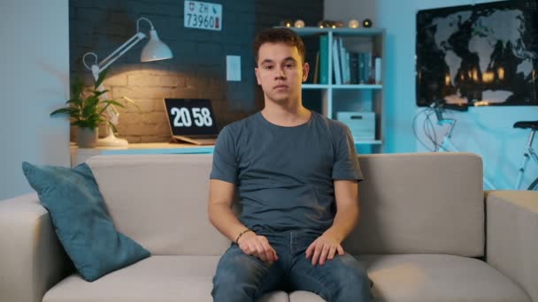 Egy aranyos tinédzser, komoly arca ül a kanapén, majd áthalad a karját nézi a kamerát — Stock videók