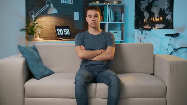 Vue de face d'un adolescent mignon avec un visage sérieux assis sur le canapé et croisant les bras en regardant la caméra . — Video