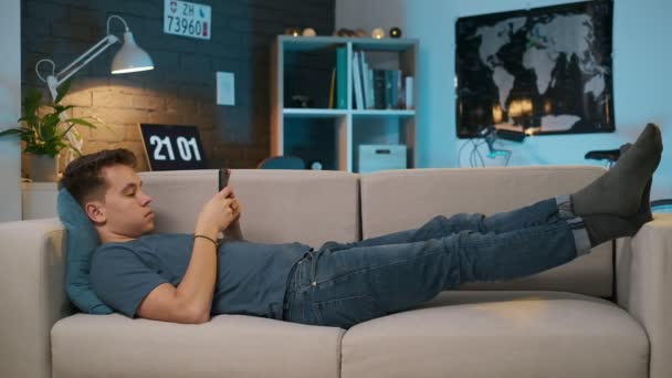 Vista lateral de um adolescente bonito deitado no sofá e jogando em seu telefone . — Vídeo de Stock