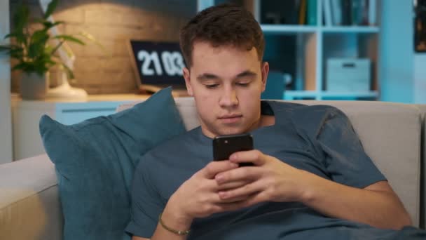 Przystojny nastoletni chłopak jest tekstylny z przyjacielem poprzez Social Media na jego inteligentny telefon siedząc na kanapie — Wideo stockowe
