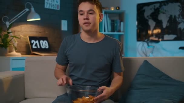 Junge sitzt auf der Couch und beginnt fernzusehen und Kartoffelchips aus der Schüssel zu essen. — Stockvideo