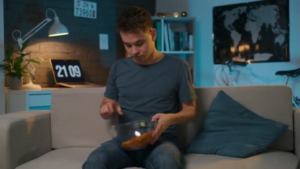 Teen boy si siede sul divano e inizia a guardare la TV attentamente mentre mangia patatine fritte — Video Stock