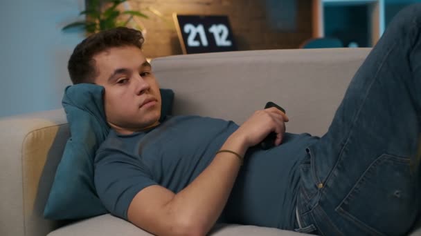 Een tiener jongen is liggend op de comfortabele bank en tv kijken, hes vervelen, zodat hij schakelt kanalen met de afstandsbediening. — Stockvideo