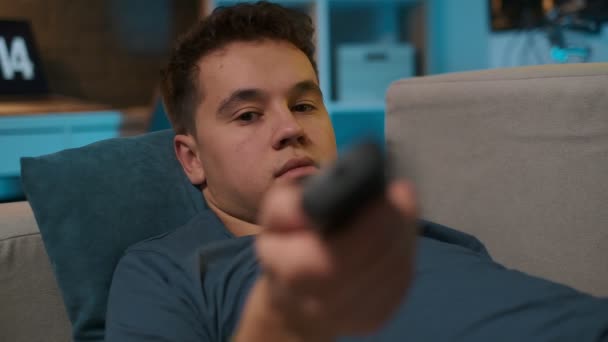 Close-up portret van een jonge kerel schakelen kanalen met de afstandsbediening terwijl liggend op de Bank en tv kijken. — Stockvideo