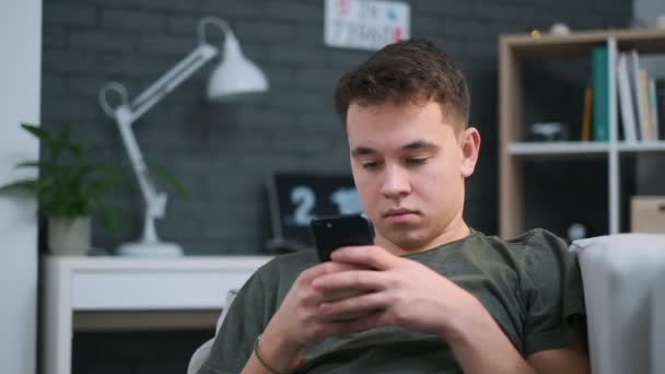 Blízký portrét mladého chlapce, který se soustředil na mobil, když odpočíval na pohovce — Stock video