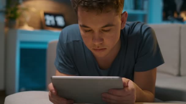 Een tiener ligt op de Bank op zijn buik en speelt op een Tablet. Close-up portret — Stockvideo