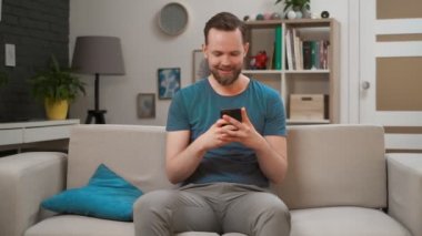 Genç, beyaz, modern cep telefonu kullanan bir adam evdeki bir koltukta oturuyor, cep telefonunu iki elinde tutuyor ve mutlu bir gülümsemeyle ekrana bakıyor.