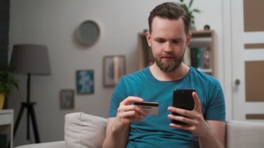 Genç beyaz adam kanepede oturan, oturma odasında kredi kartı ile online alışveriş için akıllı telefon kullanarak. Kapalı. Adam elinde kredi kartı, dijital mobil cihaz uygulaması kullanarak kolay ödeme.