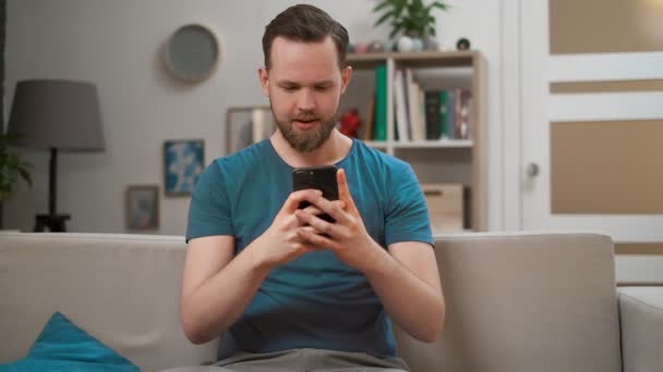 Retrato de homem caucasiano jovem usando smartphone móvel moderno sentado em um sofá em casa, segurando telefone celular em ambas as mãos olhando para a câmera com sorriso feliz satisfeito . — Vídeo de Stock