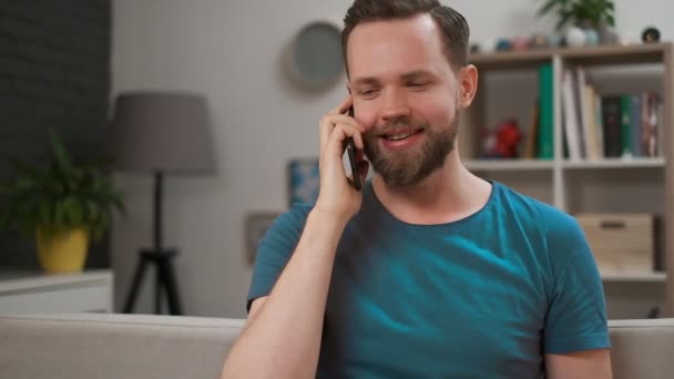 Przystojny mężczyzna rozmawia przez telefon i uśmiecha się w domu. Portret szczęśliwego Brodatego mężczyzny ubranego dorywczo rozmawiając smartfonem wewnątrz. — Wideo stockowe