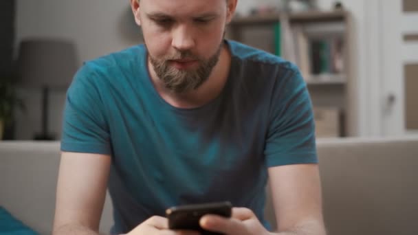 Zbliżenie kaukaski mężczyzna za pomocą nowoczesny telefon komórkowy siedzi na kanapie w domu, trzymając telefon komórkowy w obu rękach patrząc w ekran z zadowolony uśmiech zadowolony z dobrych wiadomości. — Wideo stockowe