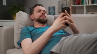 Yakışıklı bir adamın evde akıllı telefonuna yazıp mesaj atanın portresi. Erkek internettarama dijital mobil cihaz kullanarak, dinlenirken evde bağlı kalmak.