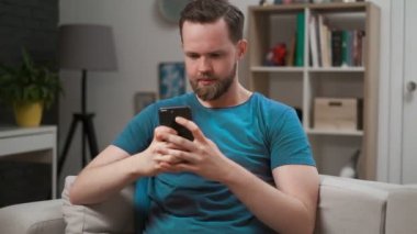 Odadaki cep telefonundaki bir uygulamada yayını kaydıran bir adamın yakın çekim görüntüsü. Evde akıllı telefonda erkek yazma ve mesajlaşma, internette gezinme, evde dinlenirken bağlı kalma.