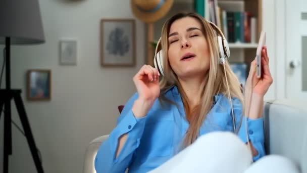 Joven chica hermosa feliz en traje azul disfrutando de la música en casa. Mujer escuchando música en auriculares desde un smartphone y bailando en la sala de estar. Cubierta, estilo de vida — Vídeos de Stock