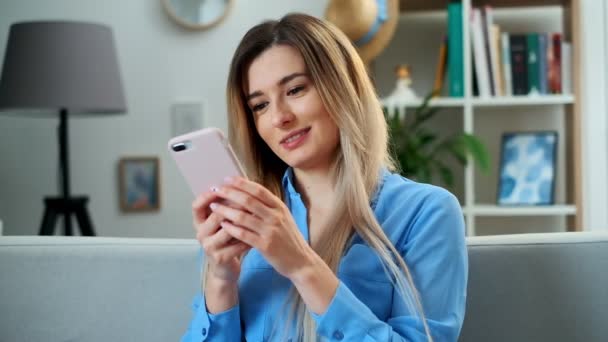 Retrato de una chica usando un dispositivo móvil navegando por Internet, permaneciendo conectada en casa disfrutando de un estilo de vida moderno. Mujer bastante joven utilizando el teléfono inteligente en el sofá, mensajes de texto mientras se relaja . — Vídeo de stock